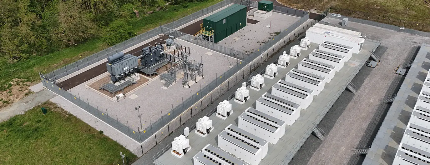 Luftaufnahme eines Stand-Alone-Batteriegroßspeichersystems mit Transformatorstation und Container-Batteriemodulen, umgeben von Sicherheitszäunen.